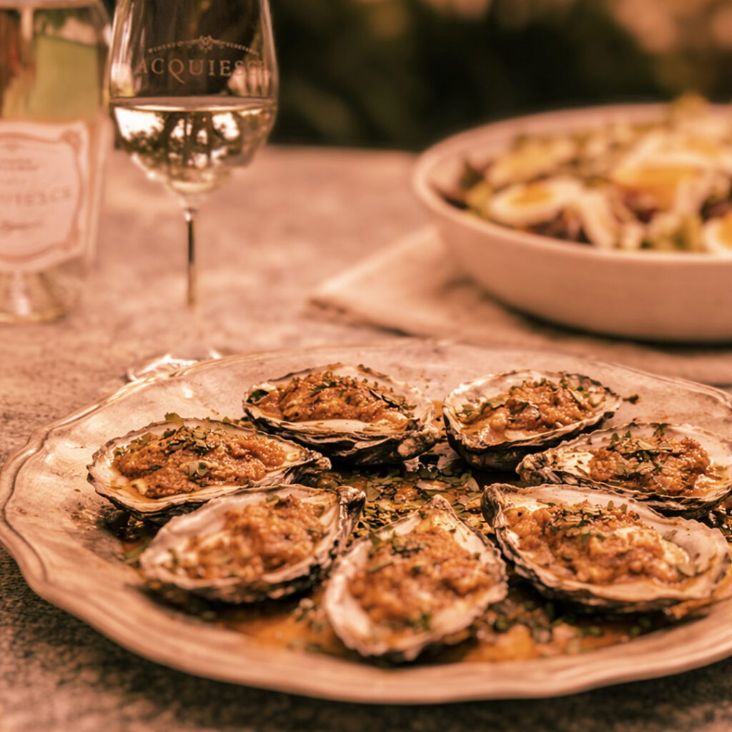 Hog Island oysters on the half shell with herbs, alongside an Acquiesce white wine bottle and glass on a stone tabletop.