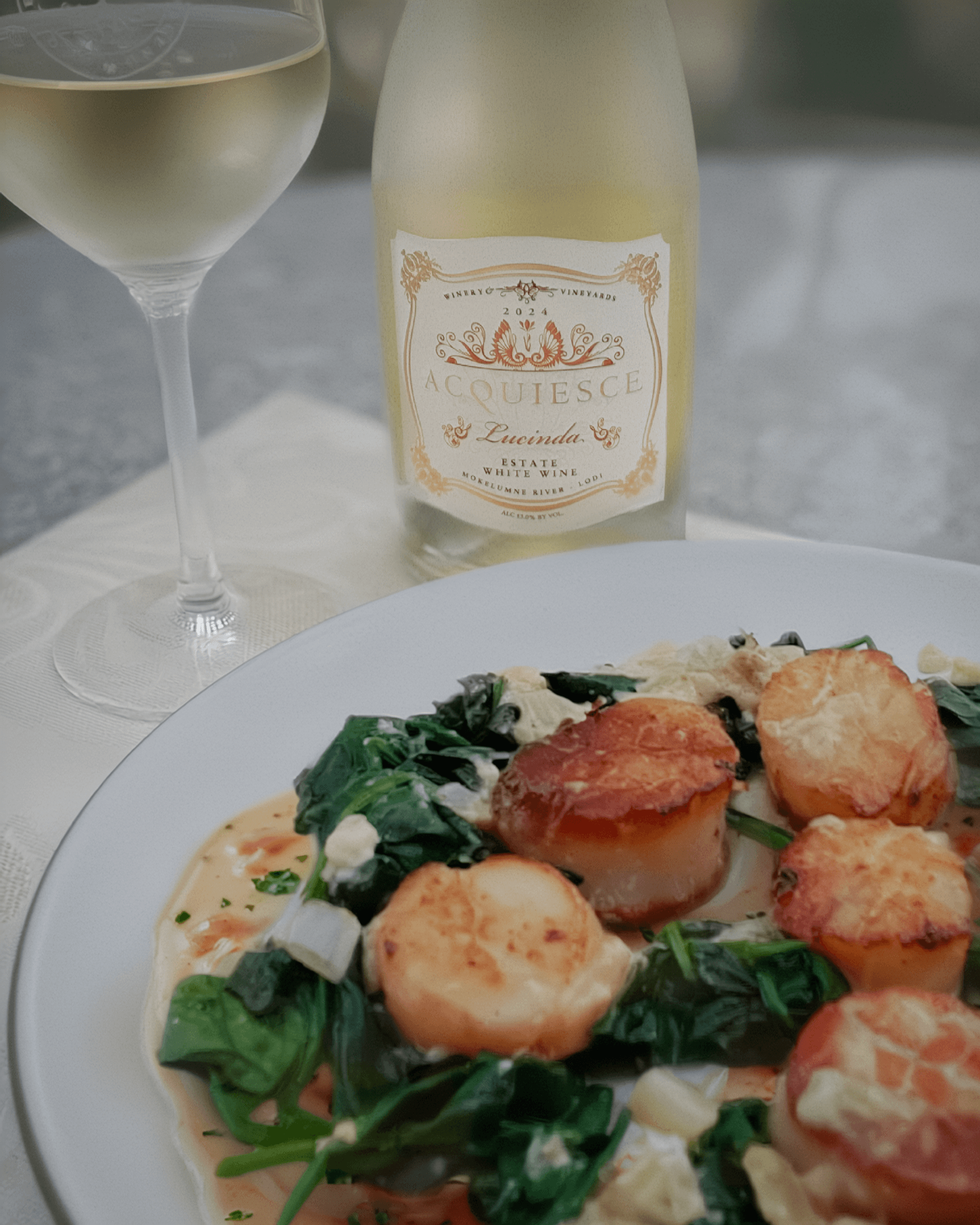 A plate of pan-seared scallops over wilted spinach with a white wine cream sauce, paired with a bottle of Acquiesce Vineyards 2024 Lucinda white wine.